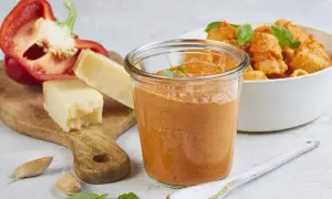 Ein Glas mit cremigem Paprika Pesto, daneben ein halber roter Paprika, Parmesan und Knoblauch. Im Hintergrund eine Schüssel Pasta mit dem Pesto.