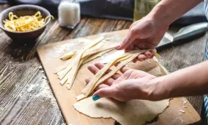 Hände formen frische Pasta-Streifen auf einem Holzbrett mit Mehl.