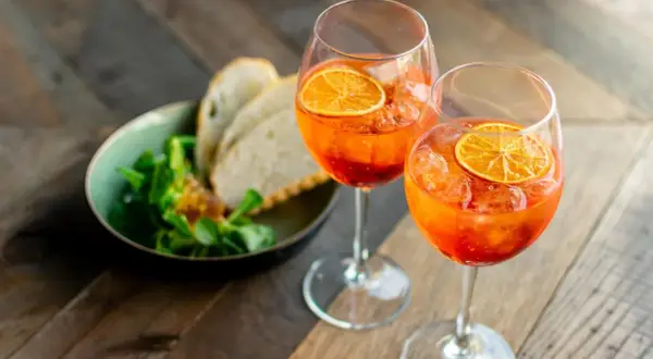 Zwei Gläser mit einem orangen Aperitif, garniert mit Orangenscheiben, daneben ein Schälchen mit Salat und Brot.