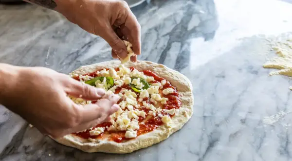 Hände belegen Pizza mit zerbröckeltem Käse auf Marmortisch.