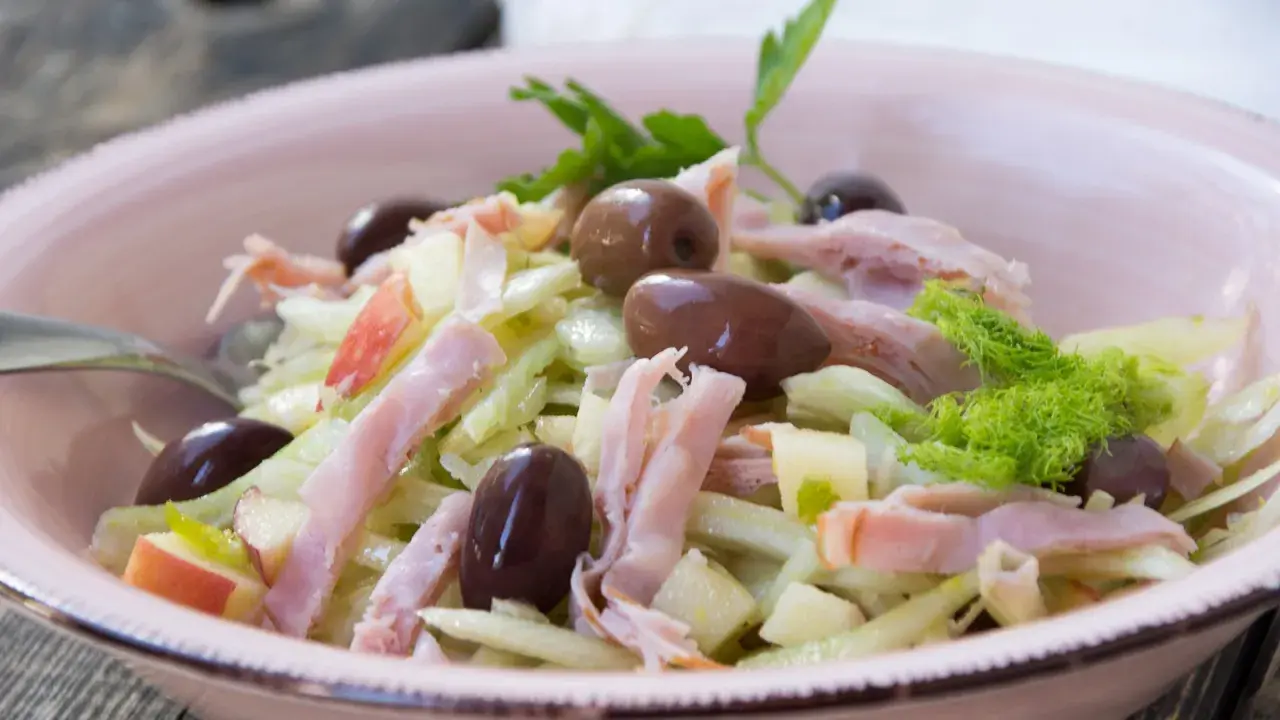 Frischer Fenchel Salat mit Apfelstücken, Schinkenstreifen und dunklen Oliven in einer rosa Schüssel.