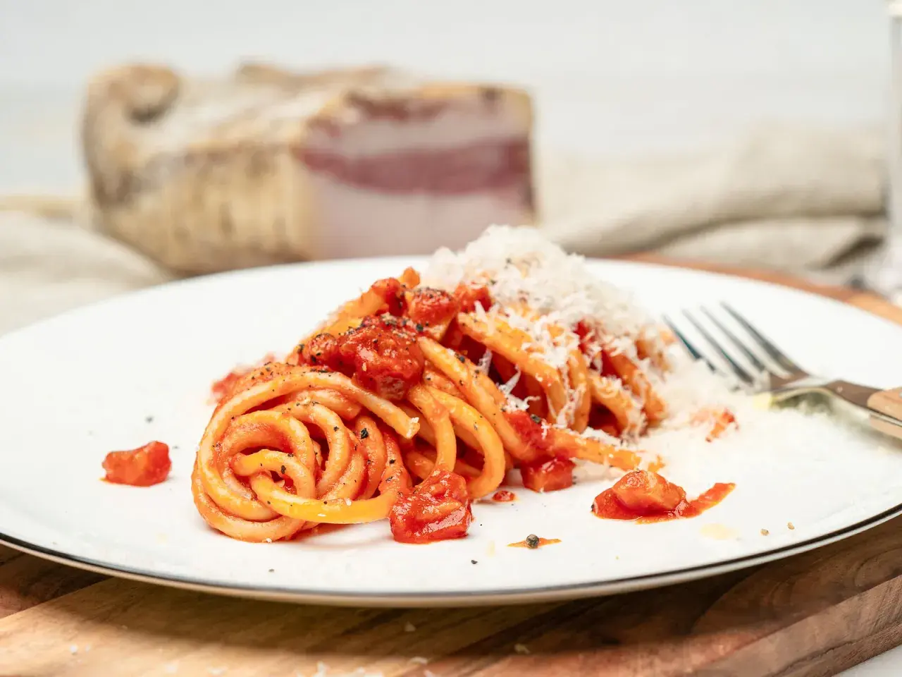 Ein Teller Pasta all'amatriciana, mit Tomaten und geriebenem Käse bestreut. Im Hintergrund ein Stück Guanciale, die Zutat für das authentische amatriciana rezept.