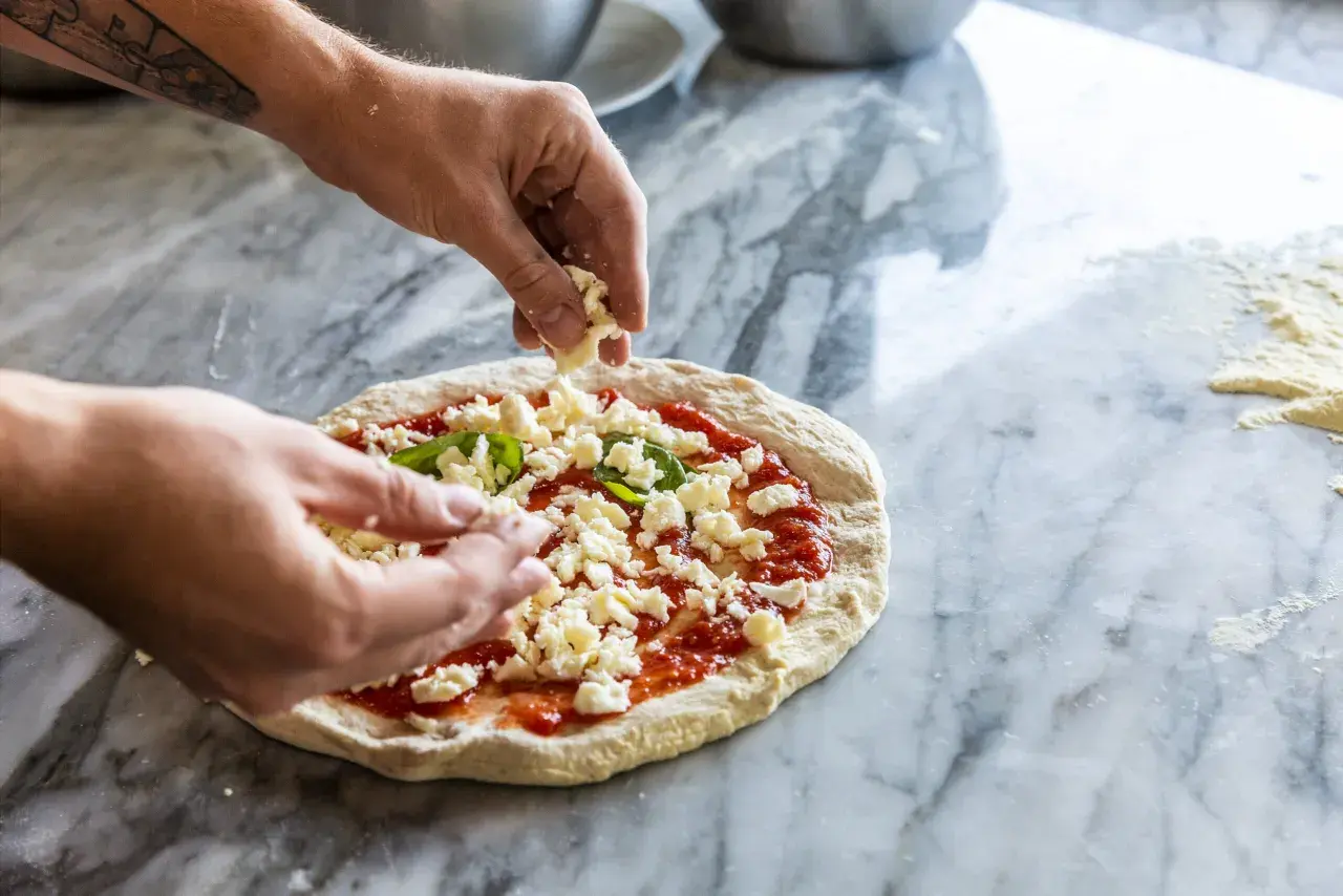 Hände belegen Pizza mit zerbröckeltem Käse auf Marmortisch.