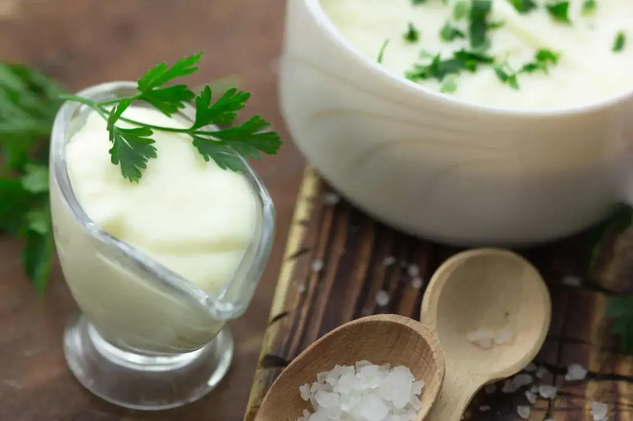 Cremige Bechamel-Sauce in einer Sauciere, garniert mit Petersilie. Daneben eine Schüssel mit der Sauce und grobes Salz auf Holzlöffeln.