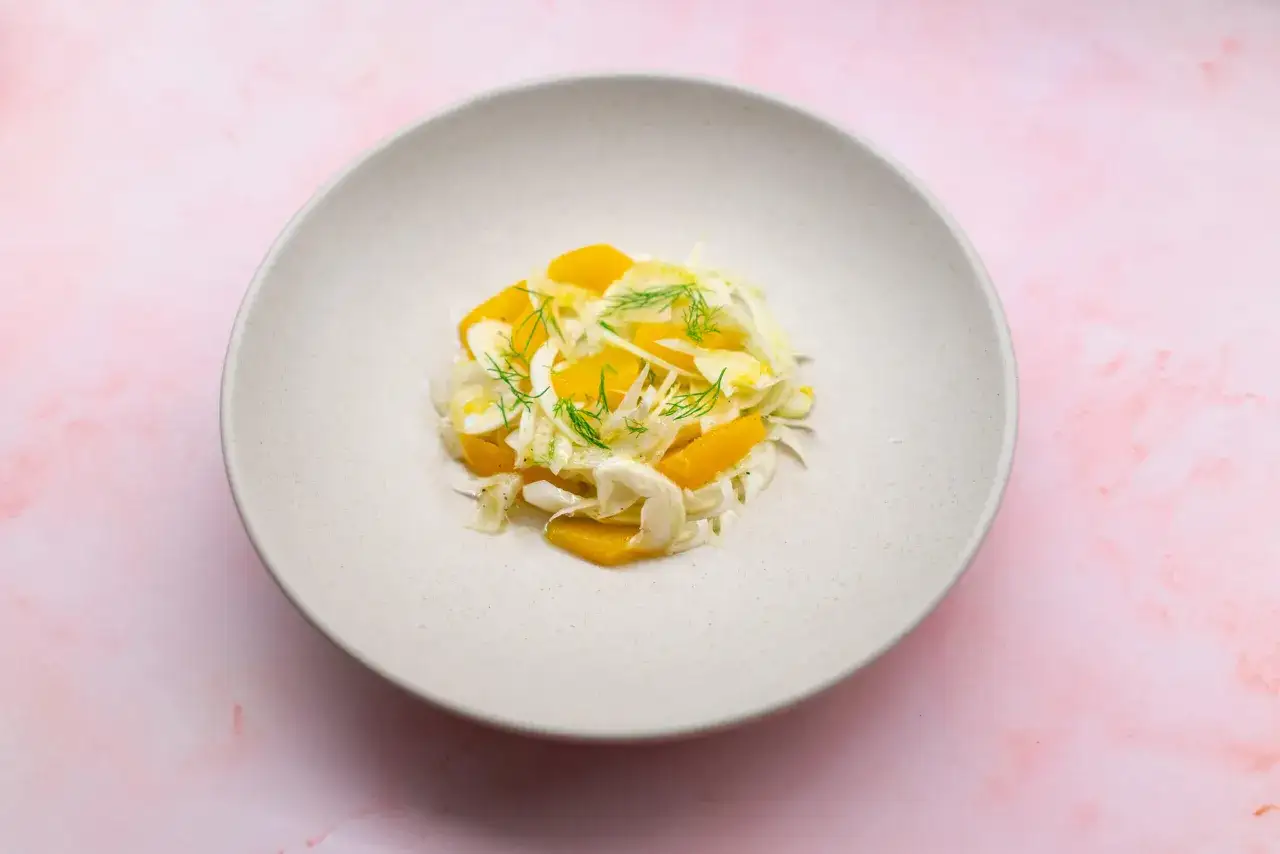 Ein leichter Fenchel Salat mit Orangenstücken und Dill garniert, serviert auf einem cremefarbenen Teller auf rosafarbenem Marmoruntergrund.