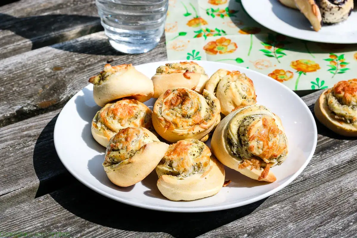 Ein Teller mit frisch gebackenen Pesto-Schnecken, garniert mit geschmolzenem Käse, steht auf einem Holztisch.