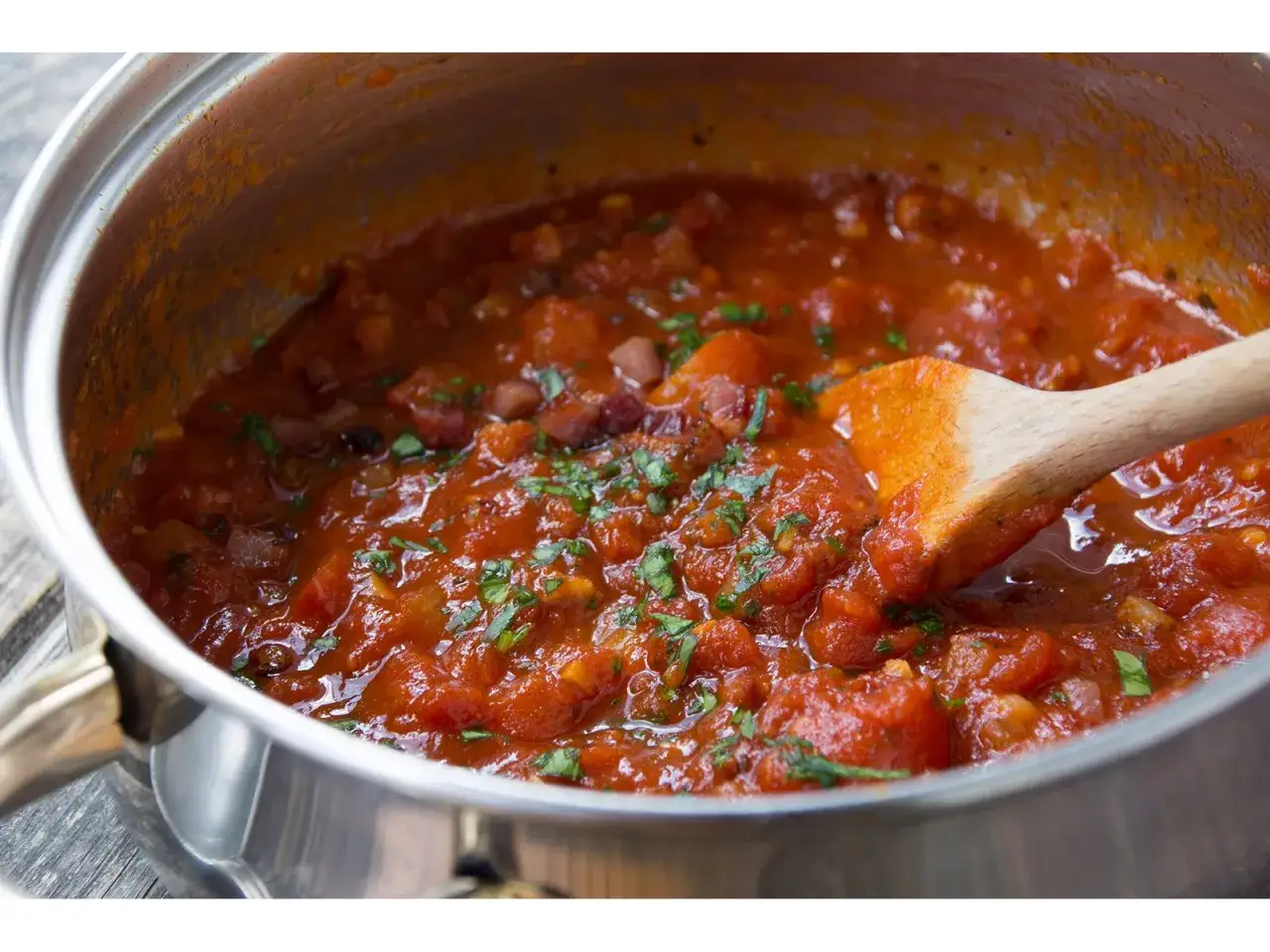 Ein Topf mit köstlicher Amatriciana-Sauce, bereit zum Servieren. Ein Holzlöffel rührt die Tomaten, Speck und Kräuter.