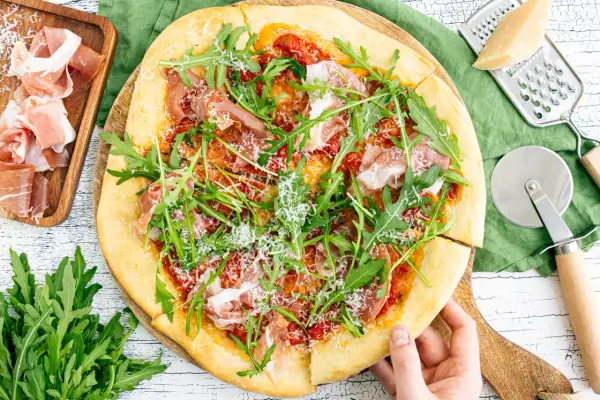 Pizza mit Rucola, Schinken und Parmesan, daneben Prosciutto, Parmesan und Pizzaschneider.