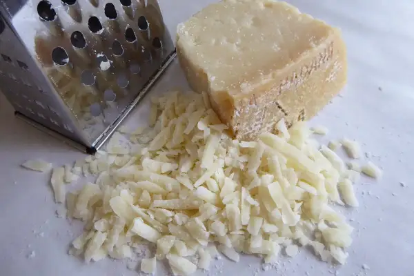Ein Stück Käse neben einem Metallreibe mit geriebenem Käse.