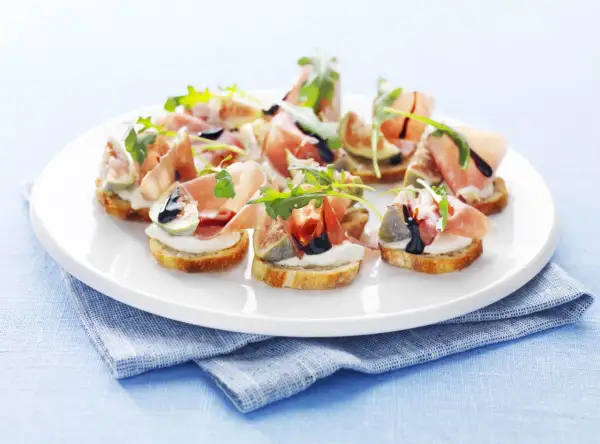 Köstliche italienische Crostini mit Prosciutto, Feigen, Mozzarella und Rucola auf einem weißen Teller.